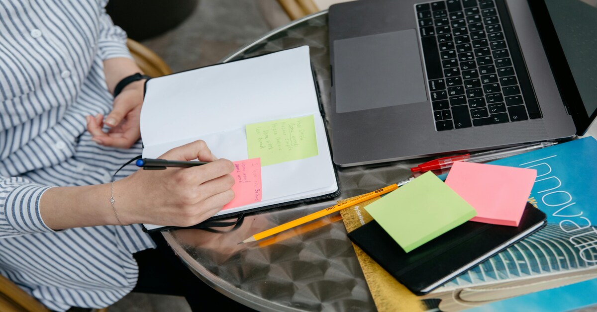 A person writes notes on colorful sticky notes next to a laptop, planning strategies to outsource content creation.