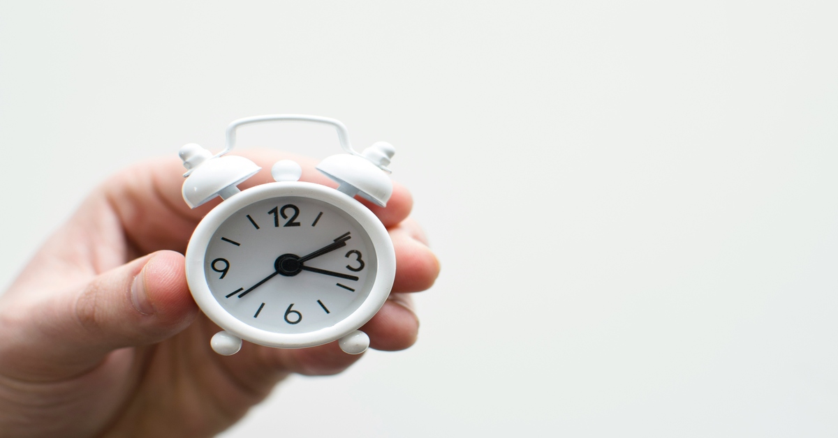 A hand holding a small white alarm clock symbolizes the importance of time management in outsourcing content creation.