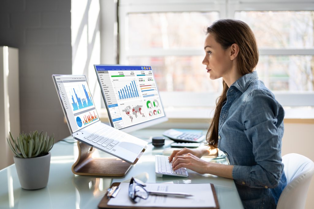 A professional woman analyzes HVAC SEO services on dual monitors displaying graphs and world data analytics in a bright office setting.