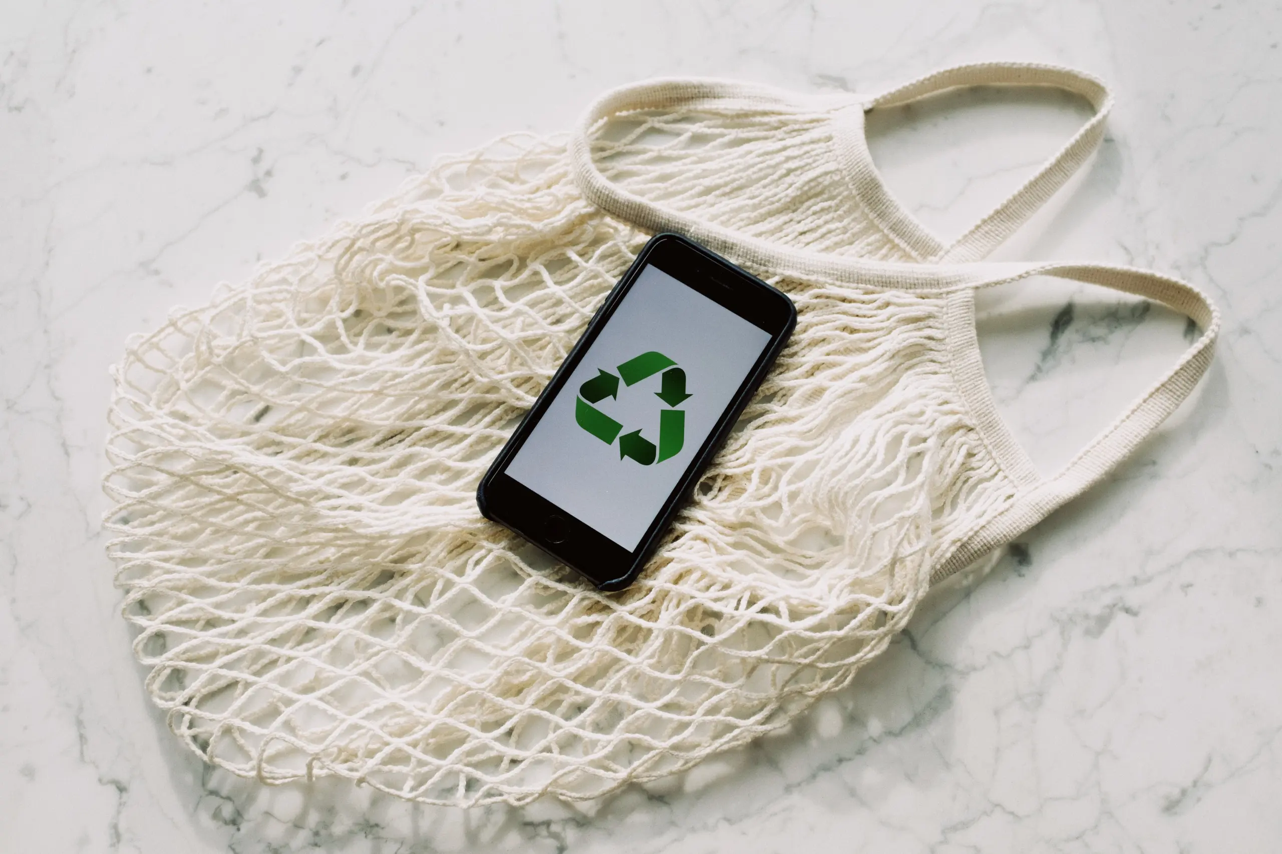 An organic shopping bag serving as a backdrop for an iPhone displaying a sustainability symbol with three green arrows on its screen. The composition conveys a message of environmentally conscious practices, combining technology and eco-friendly choices.