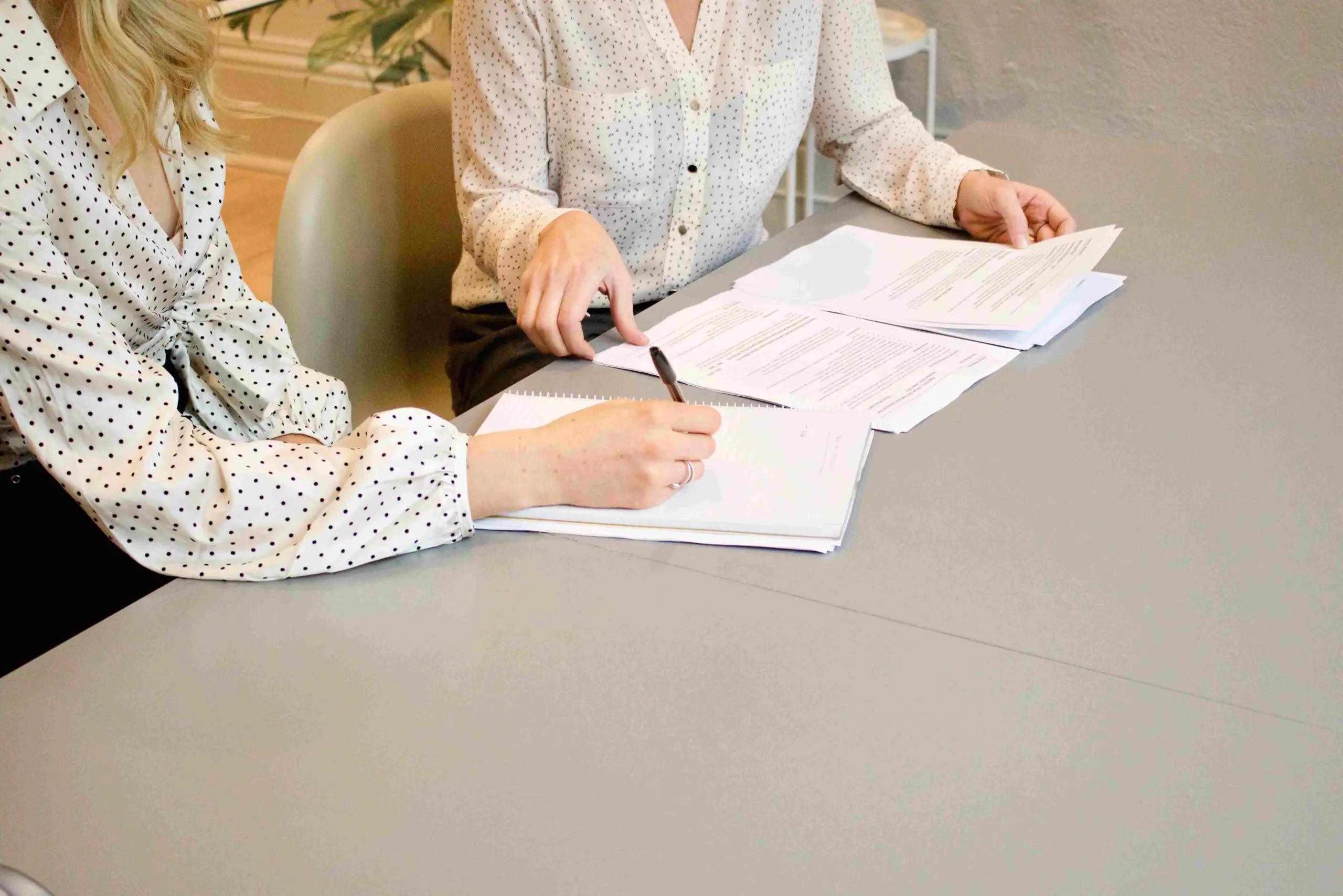 Two women wearing office attire sitting at a desk and signing contracts