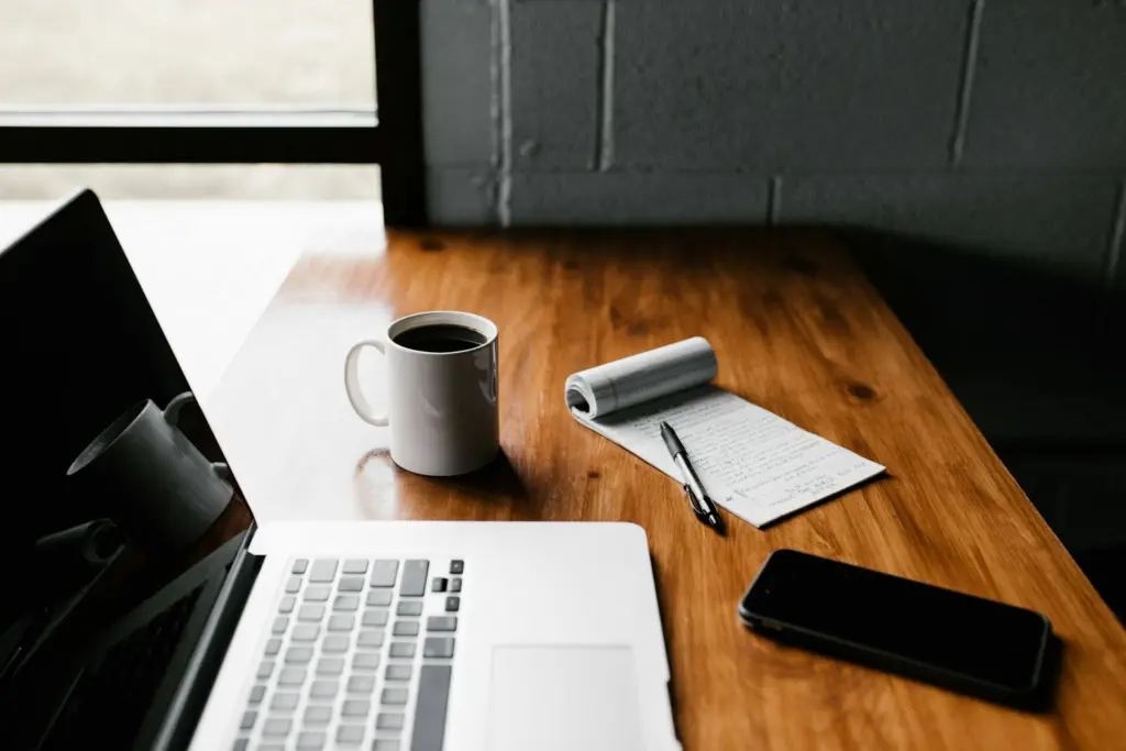 A laptop, a coffee mug, a notepad, and a smartphone on a wooden desk, essential tools for credit union advertising.