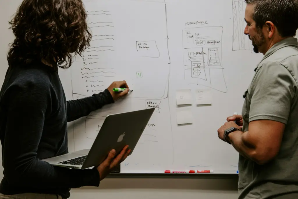 Two professionals discuss a whiteboard filled with design sketches and notes for a digital marketing strategy targeting personal injury leads.