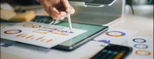 A person uses a stylus to analyze graphs and charts on paper and a digital tablet, illustrating financial data for credit union advertising.