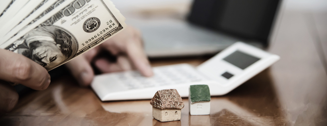 Person holding cash with a calculator and small house models, illustrating the challenges stagflation poses to financial planning.