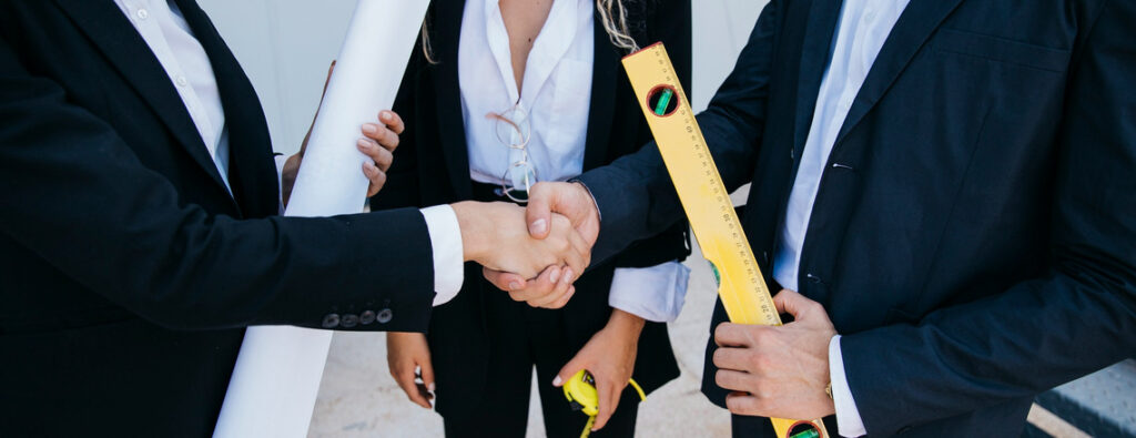 Business professionals in suits shake hands while discussing labor and construction plans, holding a measuring tool and blueprints.